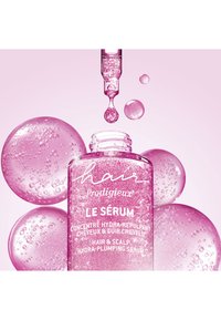 Flesje roze hydra-plumping serum, gelabeld voor haar en hoofdhuid, met een pipet die een druppel vloeistof afgeeft en roze bellen op de achtergrond.