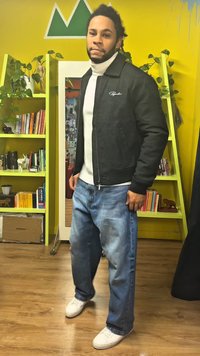 Zwarte jas met ritssluiting over een witte coltrui, gecombineerd met blauwe jeans met wijd uitlopende pijpen en witte sneakers. Binnenomgeving met gele muren en planken.