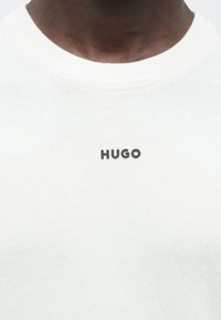 Valge puuvillasegune ümara kaelusega kampsun, millel on tekstuuriga pind ja must "HUGO" logo eesmisel küljel.