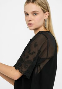 Top nero con maniche trasparenti nere e ricami intricati a motivo paisley e floreale. Design a taglio dritto, texture morbida e dettagli delicati.