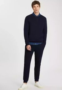 Granatowy sweter z okrągłym dekoltem, w zestawie z dżinsową koszulą i ciemnymi spodniami. Całość dopełniają białe sneakersy. Gładka tekstura na całej powierzchni.