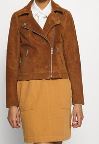 Braune Wildleder-Motorradjacke mit verkürztem Design, silbernen Reißverschlüssen und Seitentaschen, kombiniert mit einem beigen Rock und einer weißen Bluse mit einer Schleife.