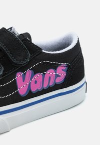 Musta kangaskengä, jossa on tarranauha, valkoinen vuoraus, valkoinen ja sininen pohja sekä rohkean pinkki "Vans"-logo sivulla kontrastivärillä ommeltuna.