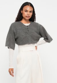 Cardigan cinza curto com mangas curtas, feito de um material peludo, apresenta um decote redondo e cinco botões pretos na frente.