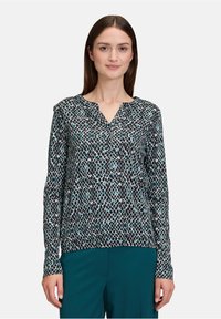 Langärmlige Bluse in Schwarz und Türkis mit Muster, die einen V-Ausschnitt, weichen Stoff und eine lockere Passform bietet. Kombiniert mit türkisfarbenen Hosen.