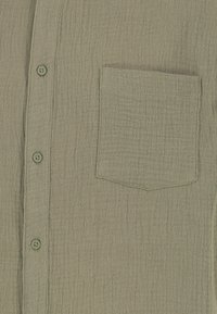 Helle grüne strukturierte Bluse mit Kragen, vorderen Knöpfen und einer einzelnen linken Brusttasche. Der Stoff ist weich und leicht zerknittert.