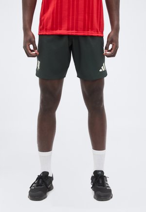 Man draagt een rood sportshirt, zwarte Adidas short met logo's, witte sokken en zwarte sportschoenen, staande tegen een effen achtergrond.