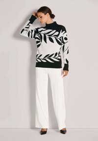 Frau im schwarz-weißen Pullover mit Blattmuster, weißen weiten Hosen und schwarzen Schuhen steht mit einer Hand an ihrem Kopf.