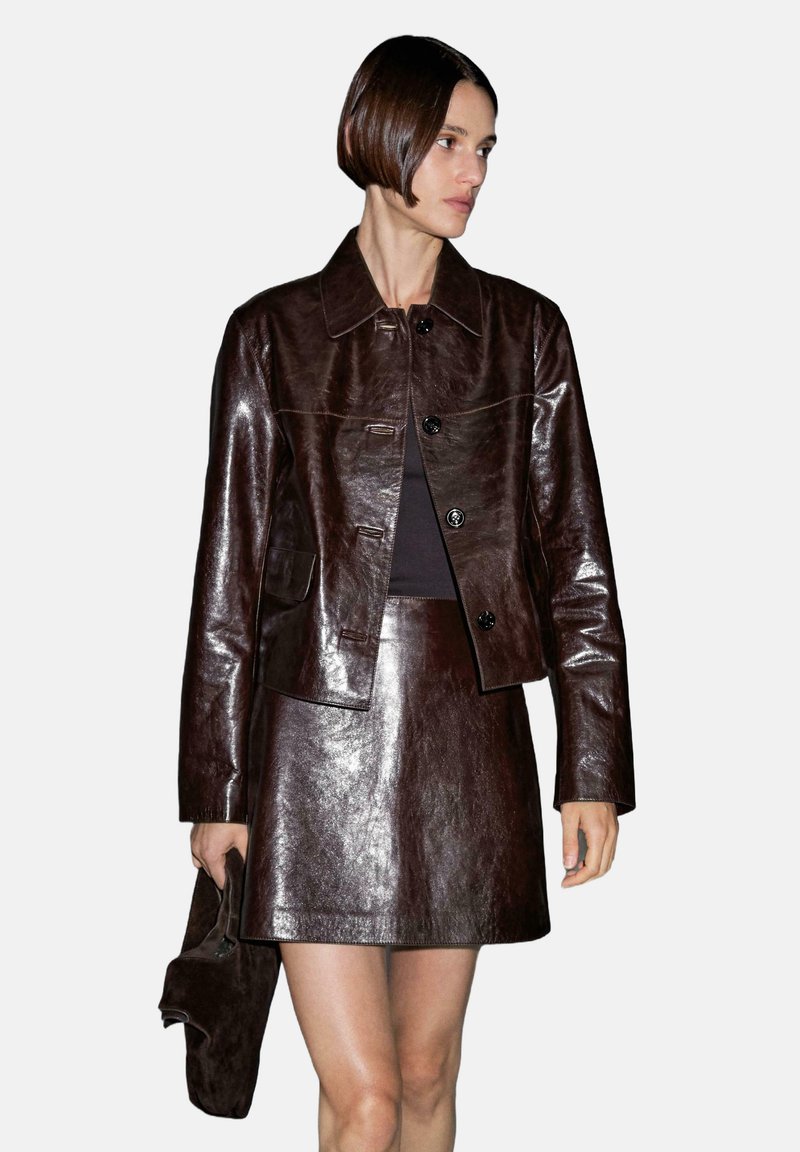 Bruine leren jas en bijpassende rok, glanzend oppervlak, strakke pasvorm, knoopsluitingen en een zak op de jas.