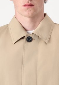 Beige jacket with a pointed collar, featuring a black button closure at the neck, smooth fabric, and visible stitching details.
