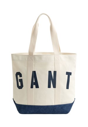 Leinwand-Tragetasche mit cremefarbenem Körper, marineblauem Boden und vertikalen Griffen, mit großen schwarzen Buchstaben "GANT" auf der Vorderseite.