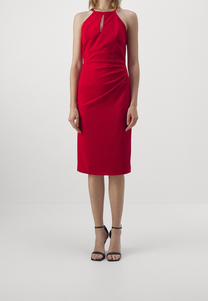 Femme portant une robe rouge sans manches, longueur genou, avec un décolleté en trou de serrure et des sandales à talons hauts à bride à la cheville noires.