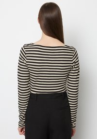 Maglietta a maniche lunghe con motivo a strisce orizzontali nere e beige; presenta una scollatura ampia e una consistenza del tessuto liscia. Pantaloni neri visibili sotto.