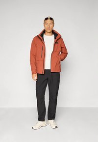 Mężczyzna w rdzawo-brązowej kurtce, białej koszuli, czarnych spodniach, białych trampkach i okularach przeciwsłonecznych na głowie, stojący na jednolitym tle.