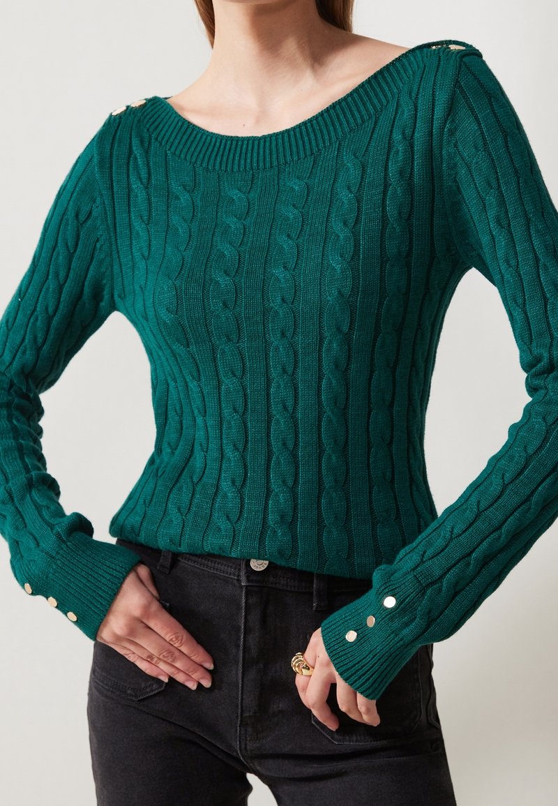 Femme portant un pull vert ajusté en maille torsadée avec des boutons argentés aux poignets et aux épaules, associé à un jean noir taille haute.