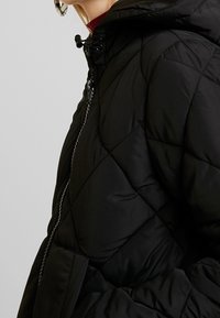 Black quilted puffer jacket with hoodie, featuring a front zipper and elastic drawstring. The fabric is smooth with a warm texture.