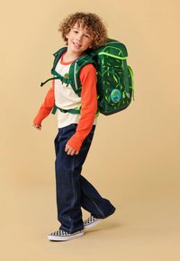 Mochila verde con patrones de rayos, correas ajustables y un parche redondo azul. Niño lleva una camiseta de manga larga naranja y jeans oscuros.