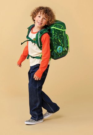 Zaino verde con motivi a saetta, spallacci regolabili e un patch rotondo blu. Il bambino indossa una maglietta arancione a maniche lunghe e jeans scuri.