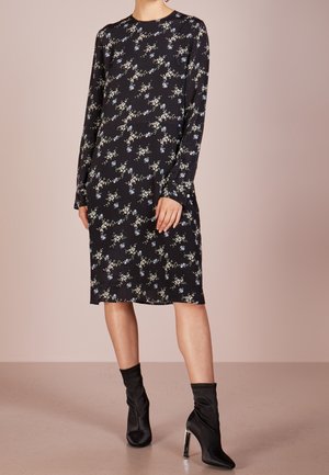 Vrouw draagt een zwarte jurk tot op de knie met een kleine witte en blauwe bloemenprint en lange mouwen, gecombineerd met zwarte enkellaarsjes met hoge hak.