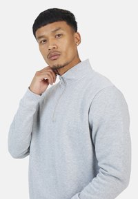 Felpa grigia chiara in pile con colletto alto e zip a quarti. Presenta maniche lunghe e una texture morbida, ideale per un abbigliamento casual.
