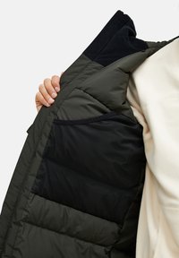 Vadderad jacka med olivgrön utsida, svart foder och en dold nätficka. Har en mjuk krage och quiltad design för isolering.