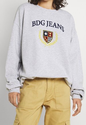 Persoon draagt een lichtgrijze BDG JEANS trui met wapen en gele lauwerkrans, gecombineerd met beige cargobroek, zichtbaar van nek tot dijen.