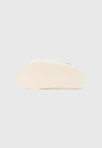 Chaussons blancs en peluche avec une semelle en caoutchouc beige texturée, présentant un logo en relief et une doublure douce pour le confort.
