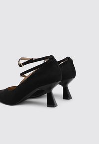 Zapatos de tacón alto de ante negro con correas en el tobillo y tacones acampanados únicos. Diseño de punta redondeada y textura suave.