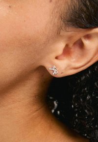 Pendiente de plata en forma de lazo, con un brillo sutil. Posicionado en la oreja de un modelo con piel texturizada y cabello rizado visible.