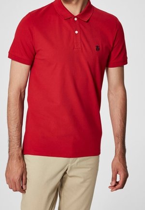 Rød polo t-shirt med korte ærmer, to-knap lukning, og et lille sort logo på venstre bryst. Båret med lyse bukser.