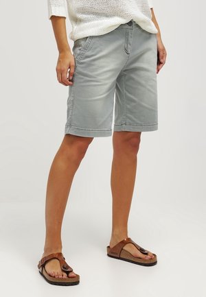 Partie inférieure du corps d'une personne portant un short gris d'une longueur jusqu'aux genoux, un pull en maille blanc et des sandales marron de style Birkenstock, debout sur un fond uni.