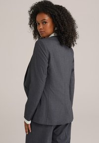 Šedý pruhovaný blazer s přiléhavým střihem, který má jednobodové zapínání, klopové límce a strukturované ramena. Texturovaná tkanina.