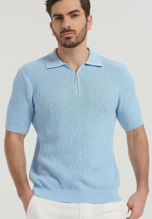 Homme portant un polo en maille bleu clair à manches courtes avec col zippé et motif texturé, associé à un pantalon blanc, posant avec les mains dans les poches.