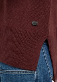 Bordeauxroter Pullover mit gerippter Textur, seitlichen Schlitzen und einem kleinen schwarzen Etikett. Kombiniert mit blauer Jeans, die einen lässigen Lagenlook zeigt.