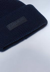 Bonnet tricoté bleu marine avec un revers, comportant une patch en tissu noir affichant le nom de la marque en lettres en relief. Apparence texturée.