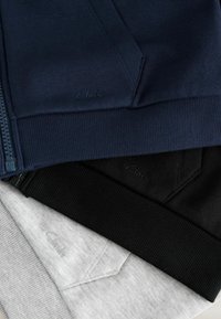 Trois sweats à capuche pliés en bleu marine, noir et gris clair, chacun avec des poignets côtelés, des poches et le logo Clarks brodé discrètement.