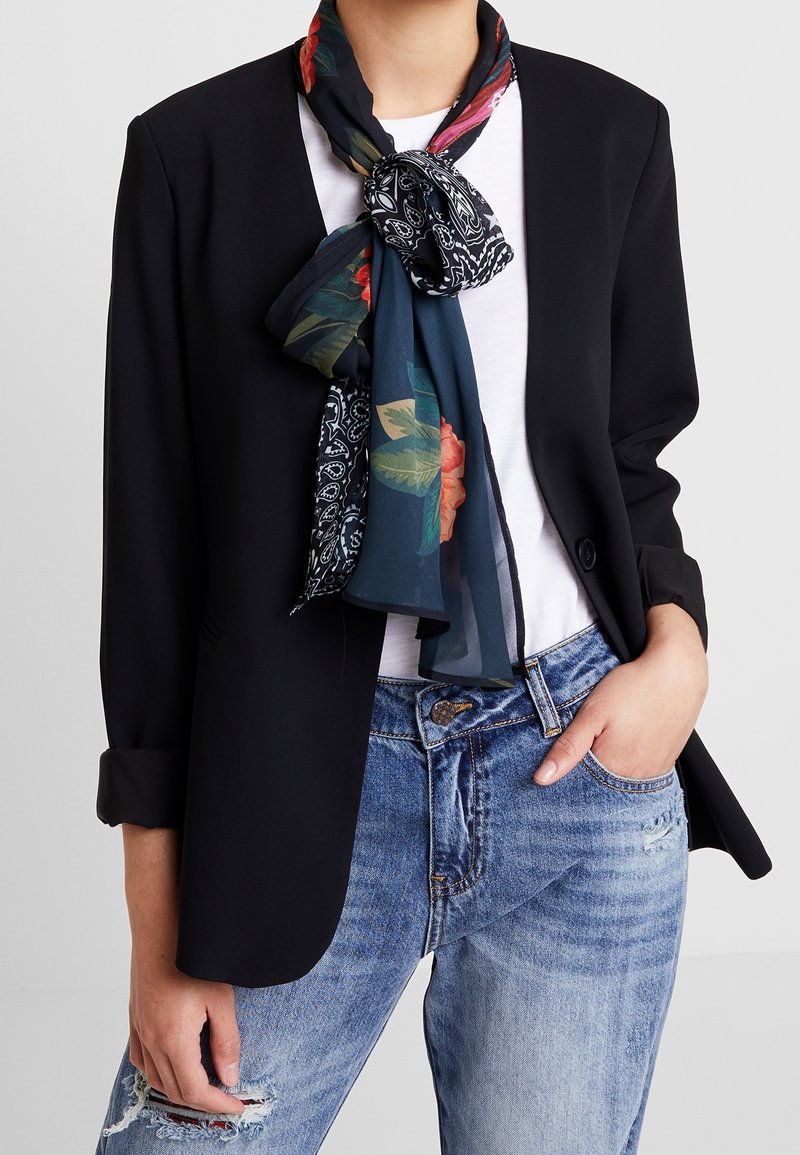 Blazer noir aux manches retroussées, associé à un t-shirt blanc et un jean bleu déchiré. Un foulard à motif floral est noué autour du cou.