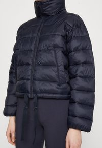 Námořnická péřová bunda s vysokým límcem, horizontálním prošíváním, předním zipem a nastavitelnými šňůrkami na spodním lemu. Měkká, lesklá látka.