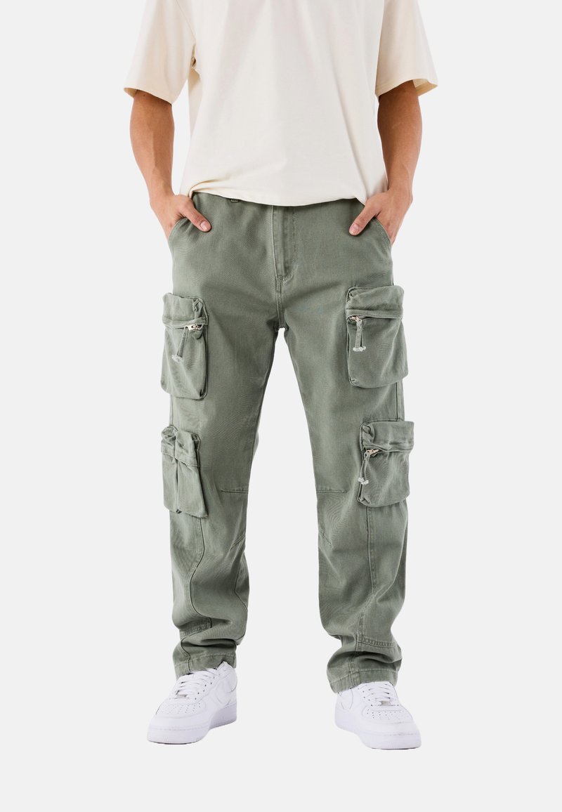 Pantalones cargo verde oliva con múltiples bolsillos con cremallera. Hechos de una tela resistente, presentan un corte holgado y pierna ajustada. Llevados con zapatillas blancas.