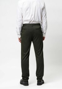 Tamsiai žali kelnės, pagamintos iš tekstūruoto audinio, su dviem užpakalinėmis kišenėmis ir siaurėjančiu siluetu, derinamos su balta ilgomis rankovėmis marškiniais.