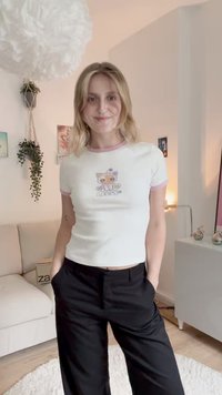 White short-sleeve t-shirt with pink ribbed collar, featuring a colorful cat graphic and text design. Worn with black pants.