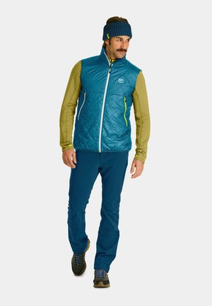 Gepolstertes Weste in Teal mit einem gesteppten Design, olivgrünes langärmeliges Oberteil mit Kapuze, blaue Hose und dunkelgrüne Wanderschuhe.