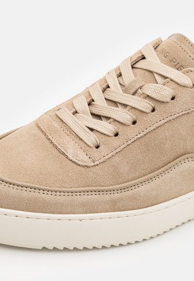 Beige suede sneaker with flat laces, padded collar, and cream rubber sole. Features textured surface and stitched detailing on the upper.