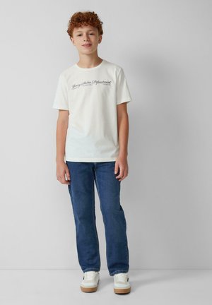 Witte katoenen t-shirt met zwarte grafische tekst, gecombineerd met blauwe spijkerbroek. De outfit is compleet met witte sneakers met een bruine zool.
