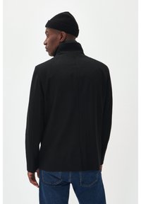 Cappotto nero con un colletto alto, design su misura e texture liscia. Indossato sopra jeans blu, abbinato a un berretto nero a coste.