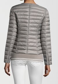 Femme portant une veste matelassée grise ajustée par-dessus un haut beige avec bordure en dentelle et un pantalon blanc, vue de dos.