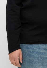 Svart langermet topp laget av mykt, teksturert materiale, kombinert med lyseblå denim. Har tettsittende mansjett og rund kant.