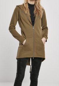 Olivově zelená mikina na zip s kapucí na šňůrku, předními kapsami, dlouhými rukávy a mírně prodlouženým zadním lemem. Hladká textura látky.