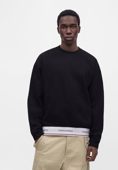 Jeune homme portant une chemise noire à manches longues par-dessus une ceinture blanche Dsquared2 et un pantalon beige, les mains dans les poches, sur un fond uni.