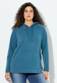 Donna con capelli ricci che indossa una felpa con cappuccio blu e jeans blu, in piedi contro uno sfondo chiaro e uniforme.